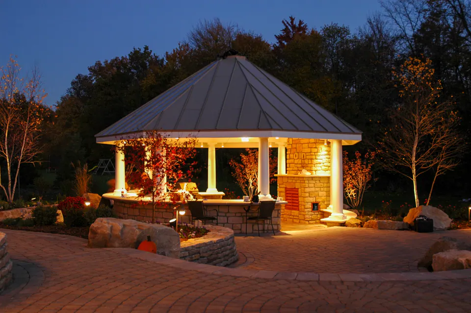 Outdoor kitchen pavilion, paver patio, stone walls, and planting lit at dusk